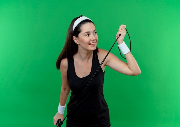 young fitness woman in headband holding skipping rope looking aside positive and happy smiling standing over green wall