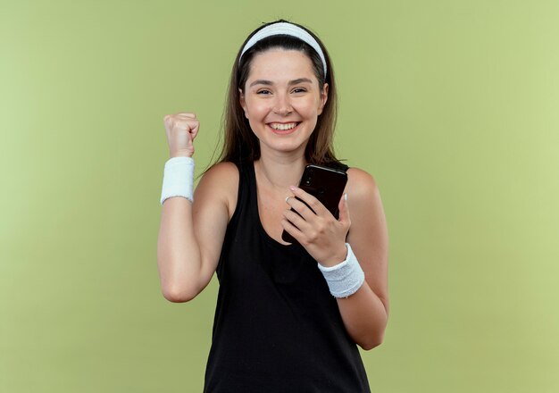 young fitness woman in headband holding smartphone clenching fist happy and excited standing over light wall