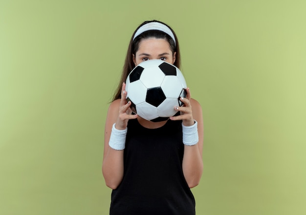 Foto gratuita giovane donna fitness in fascia tenendo il pallone da calcio che nasconde il viso dietro la palla che dà una occhiata in piedi su sfondo chiaro