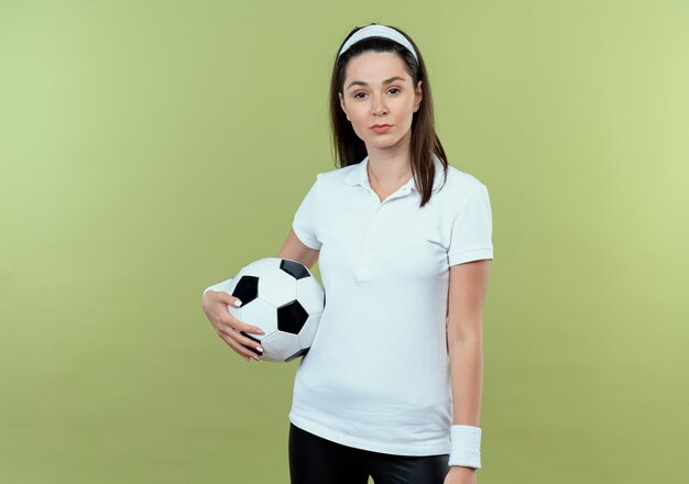 Young fitness woman in headband holding soccer ball looking at camera with serious confident expression standing over light background