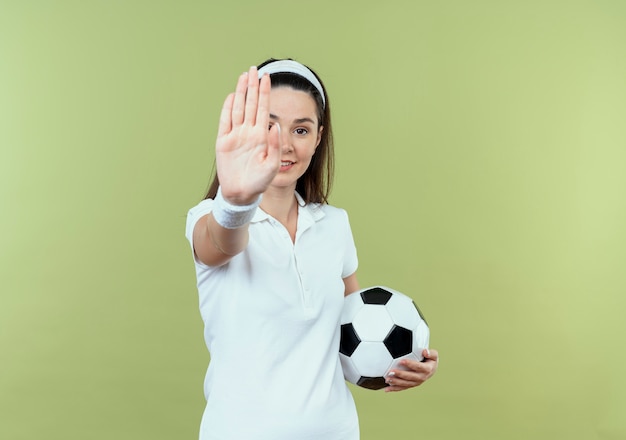 Giovane donna fitness in fascia tenendo il pallone da calcio facendo il fanale di arresto con la mano sorridente guardando la fotocamera in piedi su sfondo chiaro