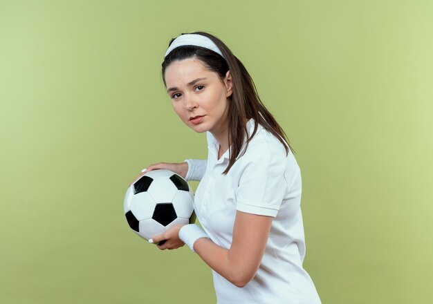 young fitness woman in headband holding soccer ball  with serious face standing over light wall