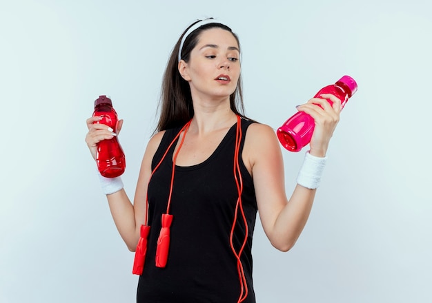 young fitness woman in headband holding two bottles of water looking confused and uncertain standing over white wall
