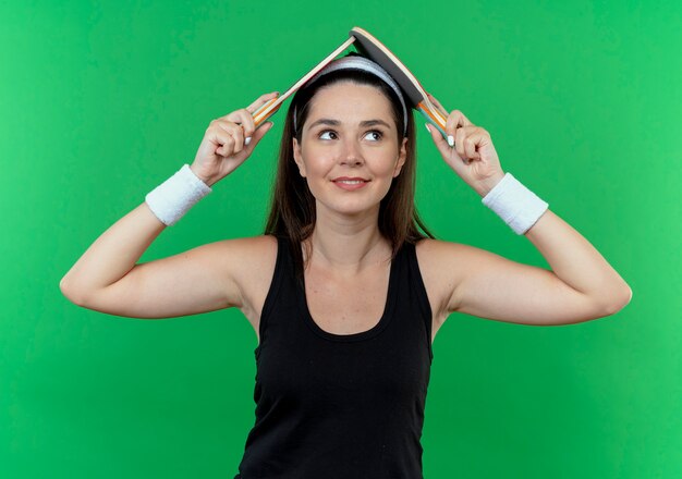 Young fitness woman in headband holding two rackets for table tennis over her head smiling standing over green background