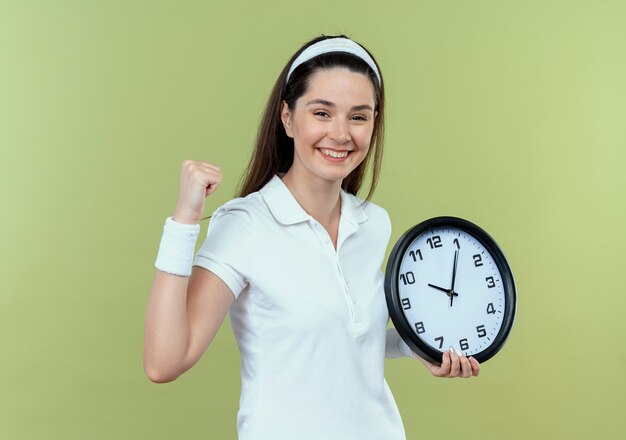 Young fitness woman in headband holding wall clock clenching fist happy and excited standing over light background