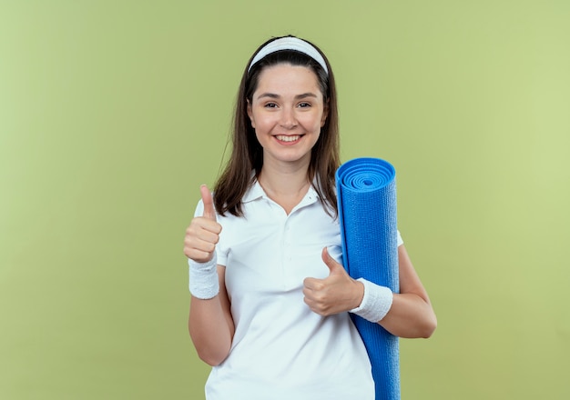 молодая фитнес-женщина в повязке на голову, держащая коврик для йоги, улыбается счастлива и позитивно показывает палец вверх, стоя над светлой стеной