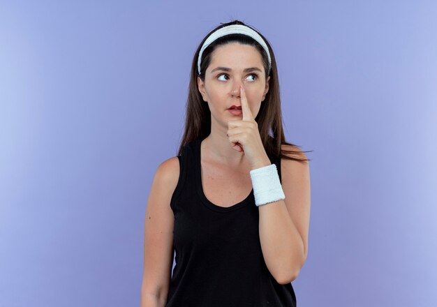 Young fitness woman in headband looking aside pointing with finger to her nose standing over blue background