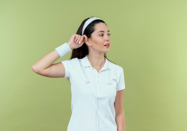 young fitness woman in headband looking aside with confident expression standing over light wall