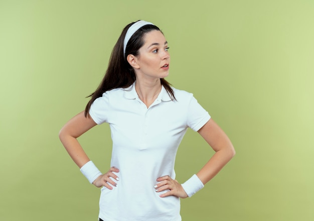 Young fitness woman in headband looking aside with confident expression with arms at hip standing over light background