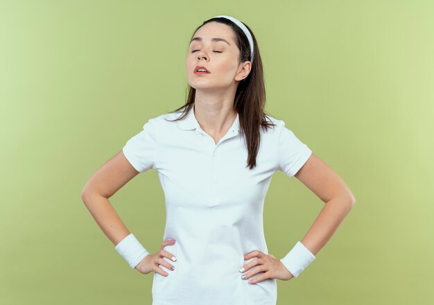 Young fitness woman in headband looking aside with confident expression with arms at hip standing over light background