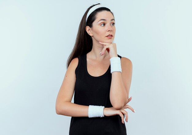 young fitness woman in headband looking aside with hand on chin thinking standing over white wall