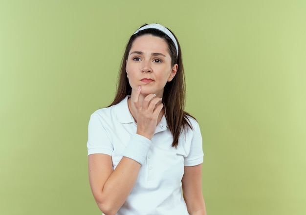 Young fitness woman in headband looking aside with hand on chin with pensive expression on face thinking standing over light background