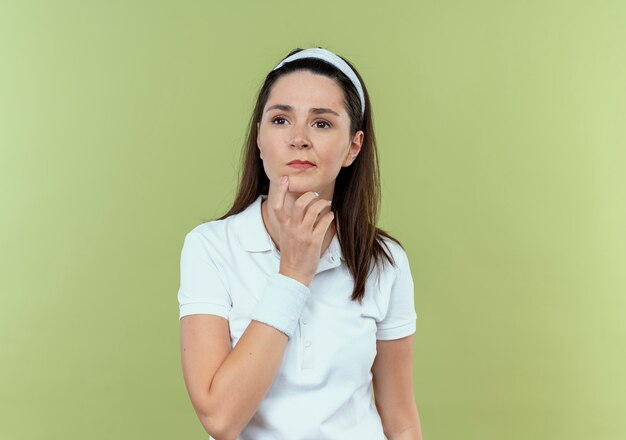 young fitness woman in headband looking aside with hand on chin with pensive expression on face thinking standing over light wall