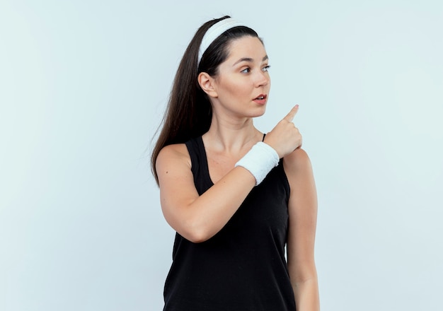 Молодая фитнес-женщина в повязке на голову смотрит в сторону с серьезным лицом, указывая пальцем ndex на что-то стоящее на белом фоне