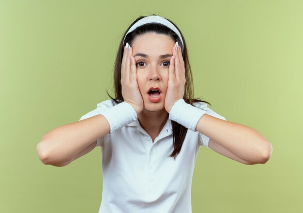 Young fitness woman in headband looking at camera being shocked standing over light background