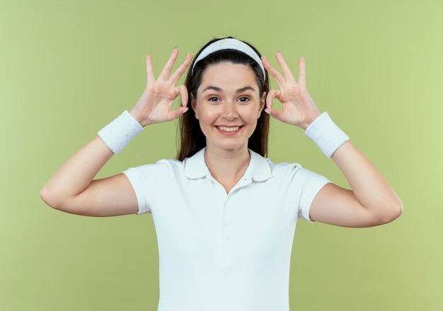 Free photo young fitness woman in headband looking at camera smiling cheerfully showing ok sign standing over light background