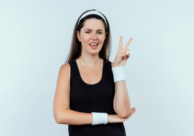 Young fitness woman in headband looking at camera smiling showing victory sign standing over white background