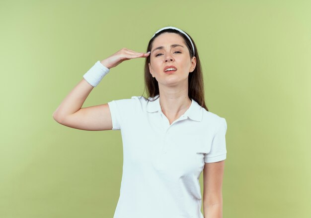 Молодая фитнес-женщина в повязке на голову смотрит в камеру с уверенным выражением лица, отдавая честь стоя на светлом фоне