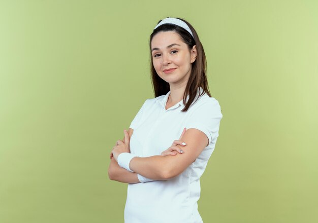 Young fitness woman in headband looking at camera with confident expression with arms crossed standing over light background