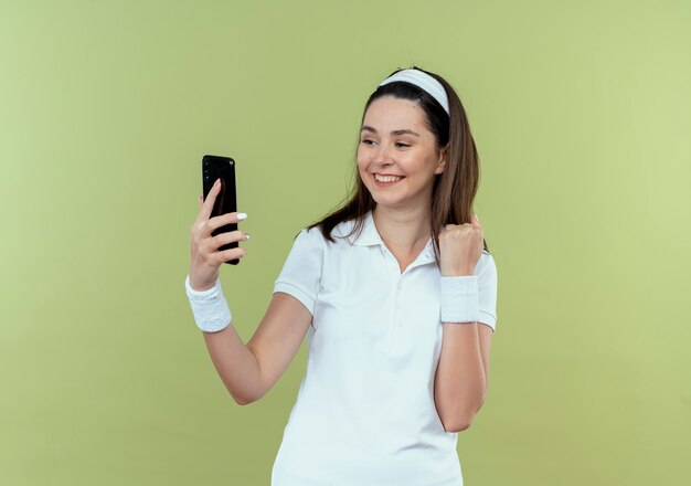 Young fitness woman in headband looking at screen of his smartphone clenching fist happy and excited standing over light wall