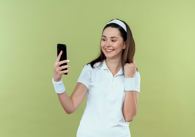 young fitness woman in headband looking at screen of his smartphone clenching fist happy and excited standing over light wall