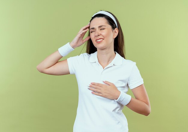 Young fitness woman in headband looking unwell touching her head having pain standing over light background