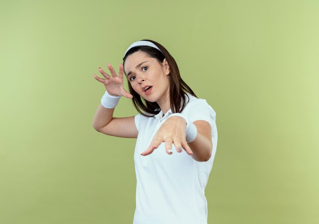 Young fitness woman in headband making defense gesture with hands looking at camera with fear expression standing over light background