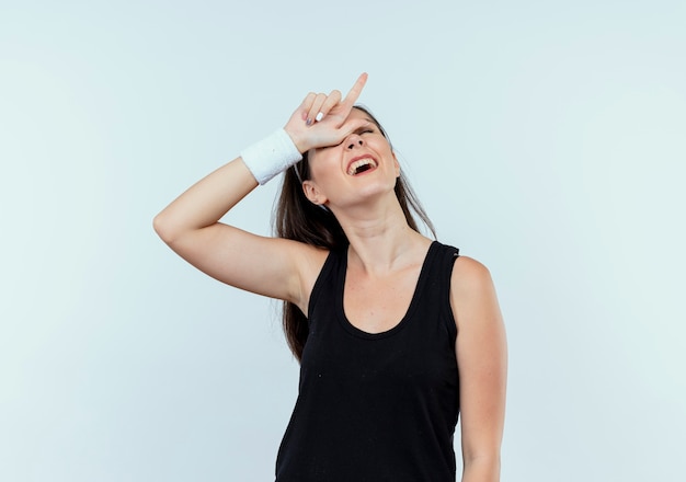 young fitness woman in headband making loser sign over her head looking confused standing over white wall