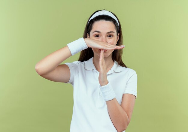 Young fitness woman in headband  making time out gesture with hands standing over light wall