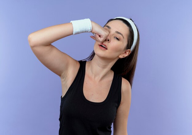молодая фитнес-женщина в повязке на голову бьет себя в лицо над синей стеной