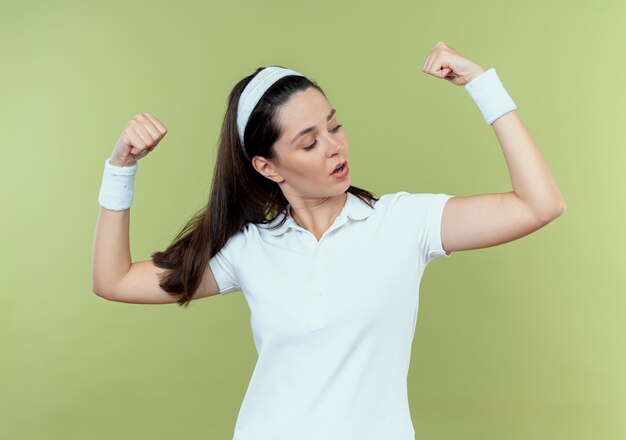 young fitness woman in headband raising fists showing biceps looking confident standing over light wall