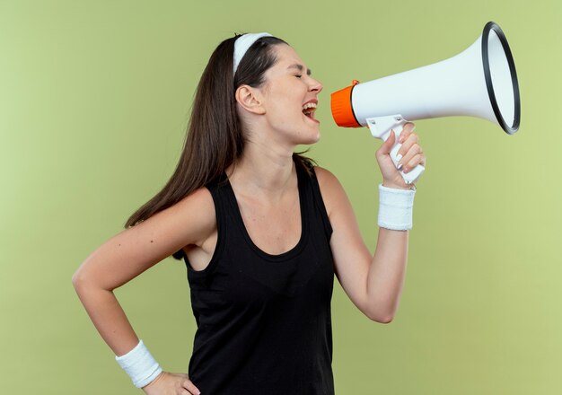 Young fitness woman in headband shouting to megaphone with aggressive expression standing over light background