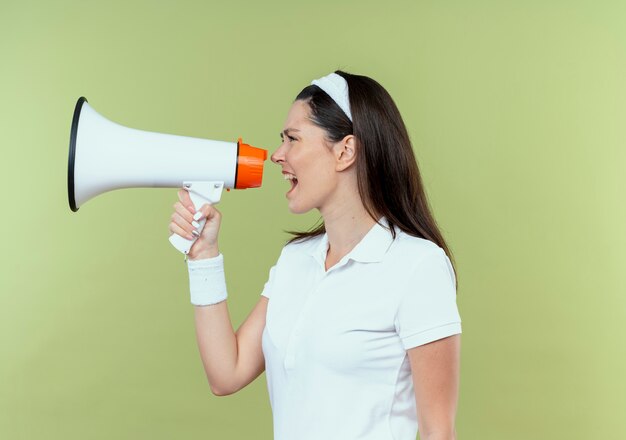 Free photo young fitness woman in headband shouting to megaphone with aggressive expression standing over light wall