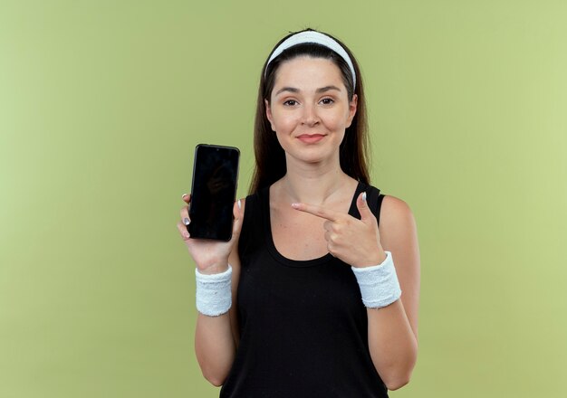 young fitness woman in headband showing smartphone pointng with finger to it smiling confident standing over light wall