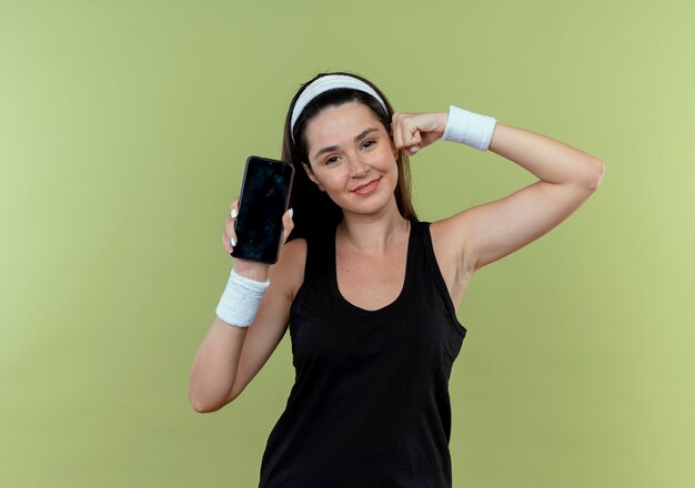 Young fitness woman in headband showing smartphone raising fist showing biceps looking confident standing over light background