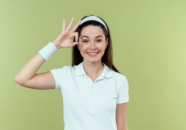 молодая фитнес-женщина в повязке на голову, весело улыбаясь, показывая знак ОК, стоящий над светлой стеной