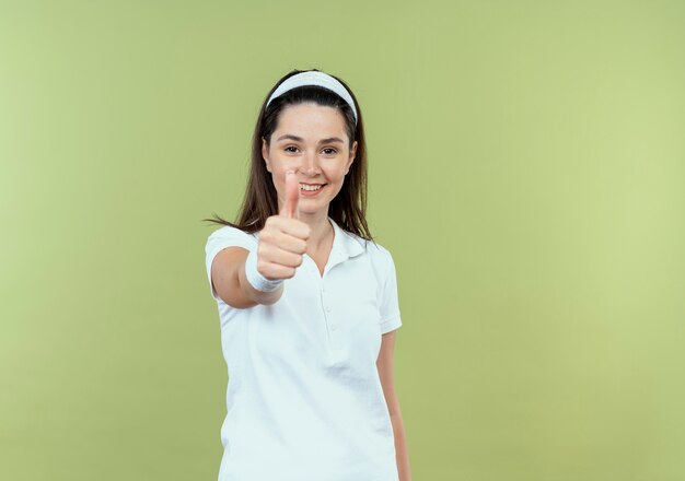 Young fitness woman in headband smiling cheerfully showing thumbs up looking at camera standing over light background