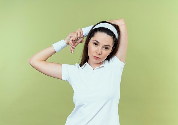 young fitness woman in headband stratching her hands looking confident standing over light wall