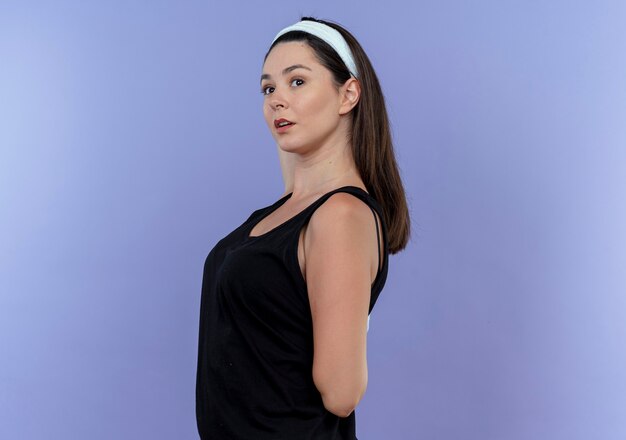 young fitness woman in headband stretching herself looking confident standing over blue wall