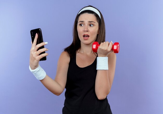 молодая фитнес-женщина в столе с повязкой на голову, держащая смартфон, тренирующийся с гантелями, выглядит смущенным, стоя у синей стены