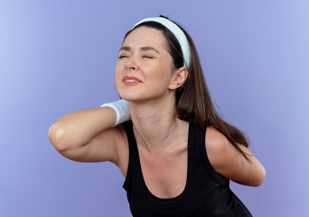 young fitness woman in headband touching her back feeling pain standing over blue wall