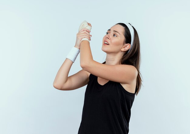 Молодая женщина фитнеса в повязке на голову, касаясь ее перевязанного запястья, чувствуя боль, стоя на белом фоне