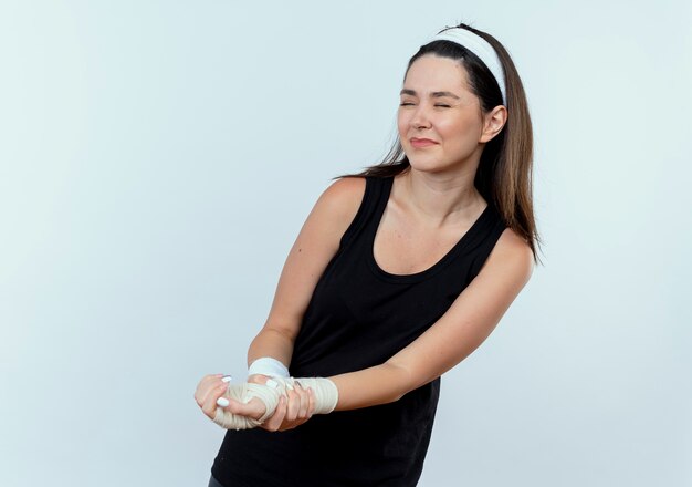 young fitness woman in headband touching her bandaged wrist feeling pain standing over white wall