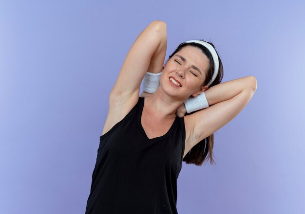 Young fitness woman in headband touching her neck suffering from pain standing over blue background