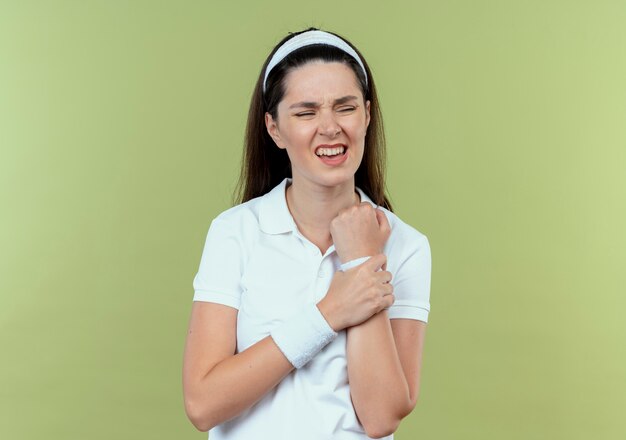 young fitness woman in headband touching her wrist looking unwell feeling pain standing over light wall