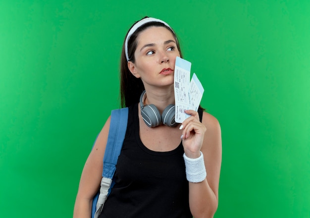 Young fitness woman in headband with backpack holding air tickets looking aside with serious face standing over green background