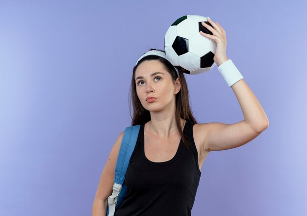 파란색 배경 위에 자신감 서 찾고 머리 위에 축구 공을 들고 배낭 머리띠에 젊은 피트 니스 여자