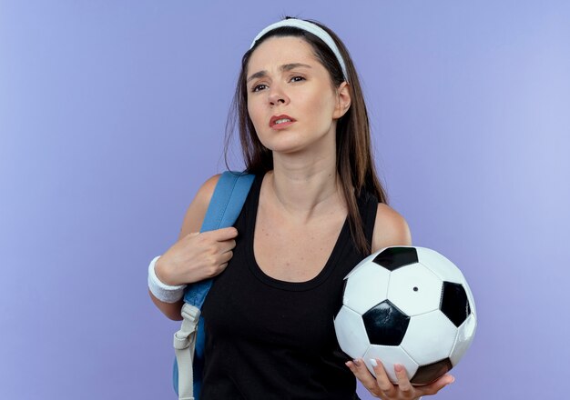молодая фитнес-женщина в повязке на голову с рюкзаком, держащая футбольный мяч, выглядит смущенной, стоя у синей стены