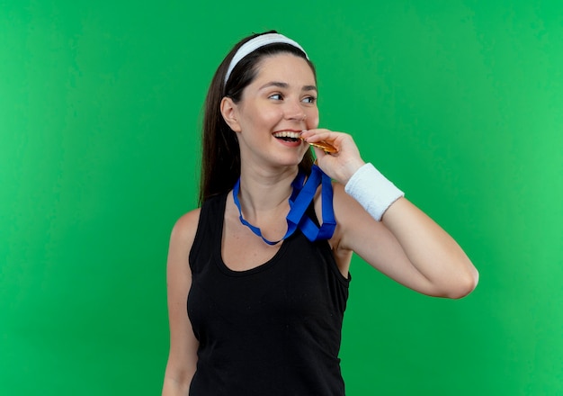 Young fitness woman in headband with gold medal around her neck biting it smiling cheerfully standing over green background