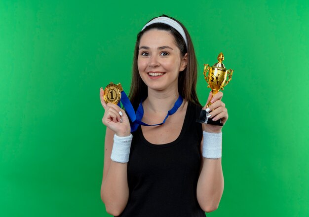 Young fitness woman in headband with gold medal around her neck holding trophy looking at camera smiling with happy face standing over green background